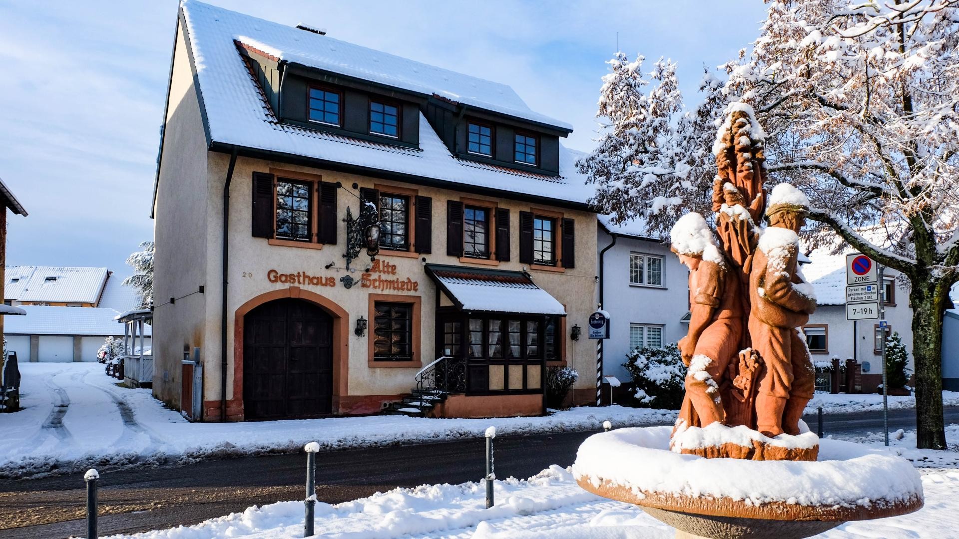 Sonnenplatz im Winter Sandsteinbrunnen mit Figurenstelen oben, die einen Fischer und einen Bauern darstellen, dahinter das alte "Gasthaus zur Schmiede"", alles schneebedeckt und sonnenbeschienen