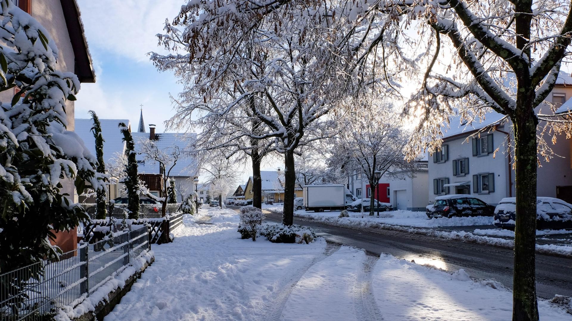 Winterstraße Straße in Rust, deren Wege, Dächer, Vorgärten und Bäume mit Schnee bedeckt sind. Von rechts blitzt die Sonne hinter den Dächern hervor.