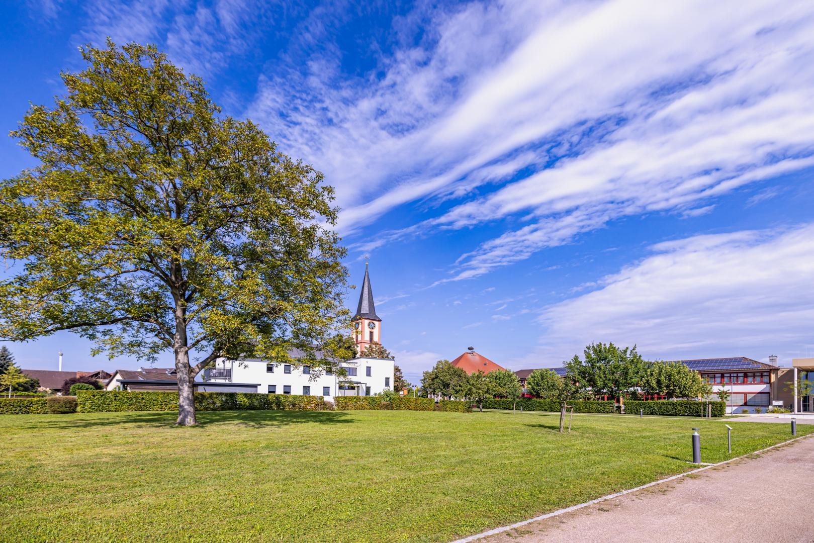 Klosgarten Rust Klosgarten, eine Wiesenfläche, im Hintergrund ein weißes, modernes Gebäude und dem KIrchtrum, darüber ein dramatischer blauer Himmel mit Wolkenfetzen