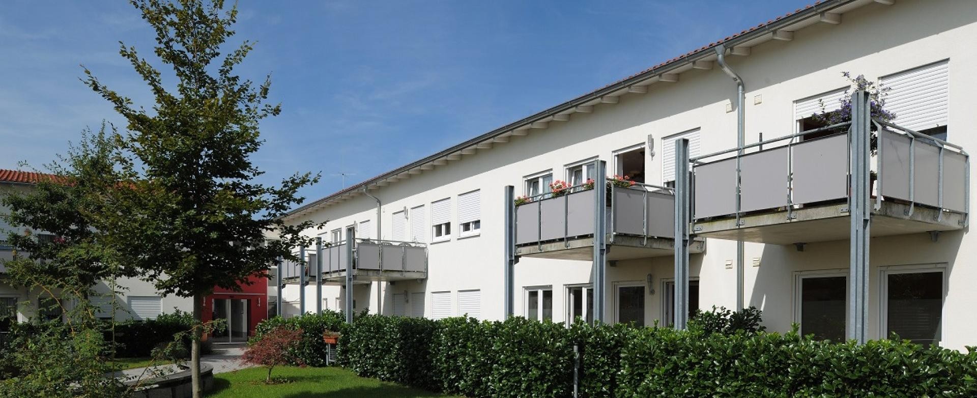 Außenfassade der Seniorenanlage "Am Klosgarten" Seniorenwohnanlage "Am Klosgarten", eine weiße Fassade mit Balkonen, darunter Terrassen, eingezäunt mit Hecken, davor eine Wiese