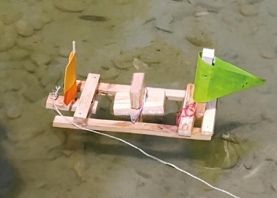 Ein Wasserrad-Schiffchen aus Holz mit einem grünen und einem orangenen Segel fährt im Wasser